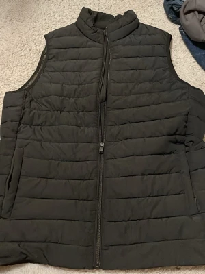Svart dunväst från Jack & Jones - Snygg svart dunväst från Jack & Jones med ståkrage och dragkedja framtill. Västen har två sidofickor och är quiltad med horisontella sömmar. Perfekt för lager-på-lager och enkel att matcha med olika outfits.