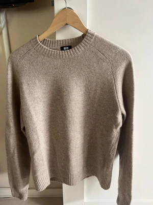 Beige stickad tröja från Uniqlo - Mysig beige stickad tröja från Uniqlo med rund halsringning och ribbade muddar vid ärmslut och nederkant. Tröjan har en enkel och stilren design som passar perfekt till lager på lager och är gjord i mjukt material.