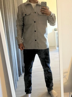 Ljusgrå overshirt med bröstfickor - Stilren ljusgrå overshirt med två stora bröstfickor och svarta knappar framtill. Skjortkrage och raka ärmar ger en clean look. Perfekt att bära över en t-shirt eller en hodie för en avslappnad men snygg stil. Materialet  är bomull och  bomullsblandning.  Han på bilden är 175 och den är lite för stor för han så skulle rekommendera 180-190 cm lång 