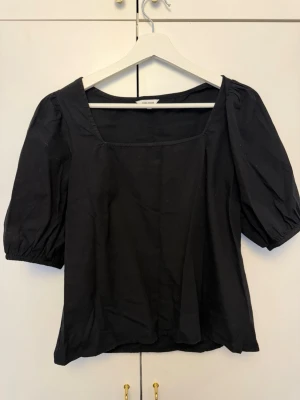 Svart blus med puffärm från Bubbleroom - Stilren svart blus från Bubbleroom med puffiga korta ärmar och fyrkantig ringning. Tillverkad i ett mjukt tyg som känns skönt mot huden. Perfekt att matcha med både jeans och kjol för en trendig look.