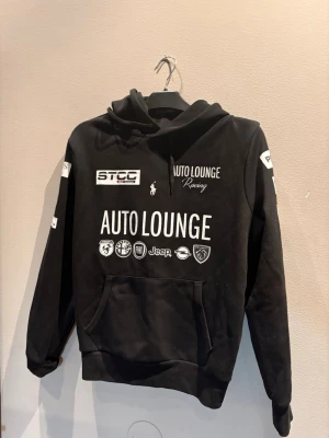 Svart Ralph Lauren hoodie med Auto Lounge-tryck - Svart hoodie med stor huva och magficka. Framsidan har flera vita tryck, bland annat 'AUTO LOUNGE', racingloggor och bilmärken som Fiat, Jeep och Peugeot. Hoodien har snörning i huvan och ribbade muddar vid ärmar och nederkant.