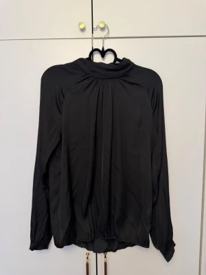 Svart blus med hög krage - Stilren svart blus med hög krage och fina rynkdetaljer fram och bak. Blusen har långärmad design och är tillverkad i ett mjukt, glansigt tyg som ger ett elegant intryck. Perfekt att matcha med både jeans och kjol.