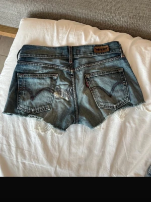 Blå jeansshorts med fransig kant - Säljer ett par blå jeansshorts med fransig, rå kant och klassiska bakfickor. Shortsen har en avslappnad passform och är perfekta för varma dagar. Snygga detaljer med slitningar och kontrastsömmar. Ikonisk denimstil som aldrig går ur tiden.