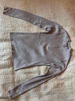 Ljusgrå ribbad stickad tröja - Säljer en ljusgrå ribbad stickad tröja med lång ärm och halv dragkedja framtill. Tröjan har en smal passform. Materialet känns mjukt och stretchigt.