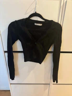 Svart ribbad croppad kofta - Snygg svart ribbad kofta med croppad passform och långa ärmar. Koftan har en knapp framtill och är tillverkad i ett stretchigt material som sitter tajt. Perfekt att styla över ett linne eller t-shirt för en trendig look.