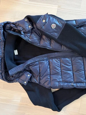 Mörkblå pufferjacka från Moncler - Snygg mörkblå pufferjacka från Moncler med stickade svarta ärmar och krage. Jackan har huva, dragkedja framtill och Moncler-logga på ena ärmen. Materialet är glansigt och quiltat, vilket ger en modern look. Perfekt för kalla vinterdagar.