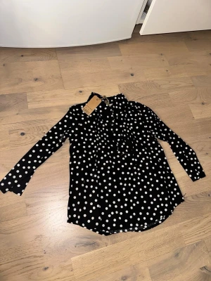 Svart prickig blus från Barpa Club - Snygg svart blus från Barpa Club med vita prickar och långärmad design. Blusen har en klassisk krage och knappar framtill. Perfekt för dig som gillar retrovibbar och lekfulla mönster. Materialet känns lätt och luftigt.Svart prickig blus från Barpa Club