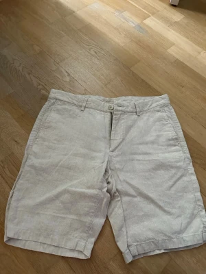 Beiga linneshorts stl 42 mycket fint skick  - Snygga beiga shorts i linne med klassisk passform och knappstängning framtill. Shortsen har bälteshällor, sidofickor och två bakfickor med knapp. Perfekta för varma dagar och ger en avslappnad men stilren look.