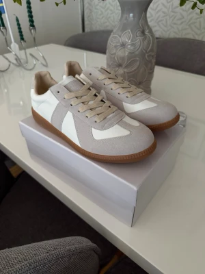 Gråa Maison Margela Gats  - Stilrena sneakers i beige mocka och vitt skinn med klassisk brun gummisula. Låga i modellen med matchande beige snören och snygga detaljer. Perfekta för dig som gillar minimalistisk och tidlös stil.