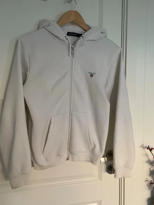 Vit zip-hoodie från Gant  - Vit hoodie med dragkedja från Gant. Tröjan har huva med dragsko, två fickor framtill och Gant-logga broderad på bröstet. Perfekt för en avslappnad och stilren look.