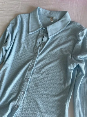 Ljusblå skjorta - Säljer en ljusblå skjorta med ribbad struktur och långa ärmar. Skjortan har klassisk krage och knäppning framtill med matchande knappar. Perfekt för en stilren och fräsch look.