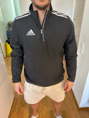 Svart Adidas träningsjacka - Säljer en svart träningsjacka från Adidas med vit dragkedja och vita ränder på axlarna. Jackan har hög krage, lång ärm och Adidas-logga på bröstet. Perfekt för träning eller uppvärmning. Materialet är lätt och andas bra. Skulle säga att den är typ small. 