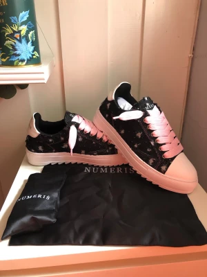Numeris sneakers med rosa snören - Snygga sneakers från Numeris med svart och vit mönstrad ovandel, vita tåpartier och rosa breda skosnören. Skorna har en chunky vit sula och detaljer i ljusrosa på hälen. Tillverkade i konstläder med textilfoder. Perfekta för dig som vill sticka ut.