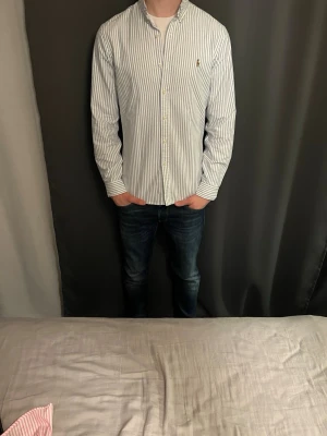 Vit och blå randig skjorta - Klassisk vit skjorta med tunna blå ränder och knappar framtill. Skjortan har lång ärm och normal passform, perfekt för en clean och stilren look. Tillverkad i bomull för en skön känsla hela dagen.