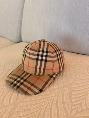 Burberry Keps - Säljer en klassisk beige keps från Burberry med det ikoniska rutmönstret i svart, vitt och rött. Kepsen har böjd skärm och justerbart spänne bak samt en svart patch med texten 'Burberry London England'. Perfekt accessoar för en trendig look.