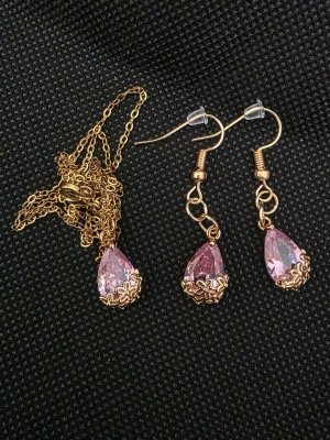 Set med halsband och örhängen i guldton - Säljer ett matchande set med halsband och örhängen i guldton med droppformade rosa stenar. Kedjan är tunn och smyckena har fina detaljer i metall runt stenarna. Perfekt för dig som gillar glittriga accessoarer och vill sticka ut.