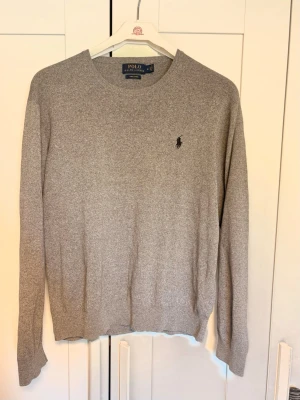 Grå stickad tröja från Polo Ralph Lauren - Klassisk grå stickad tröja från Polo Ralph Lauren med rund halsringning och diskret broderad logga på bröstet. Tröjan har långa ärmar och ribbade muddar vid ärmslut och nederkant. Perfekt till lager på lager och enkel att matcha.