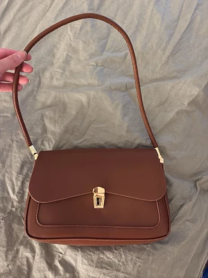 Brun axelremsväska i läderlook - Snygg brun handväska med axelrem och guldfärgade detaljer. Väskan har ett klassiskt, fyrkantigt snitt och stängs med ett elegant guldfärgat spänne. Invändigt finns ett fack med dragkedja för extra förvaring. Perfekt för dig som gillar stilrena accessoarer.