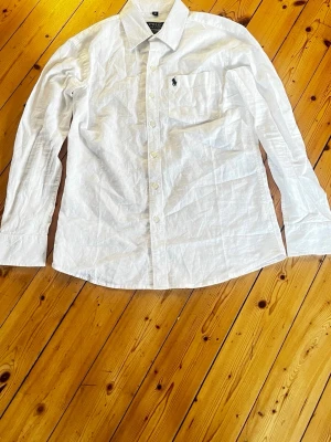 Vit skjorta från Polo Ralph Lauren - Klassisk vit skjorta från Polo Ralph Lauren med lång ärm och knappstängning framtill. Skjortan har en bröstficka med den ikoniska broderade loggan. Tillverkad i bomull och har en tidlös, normal passform.