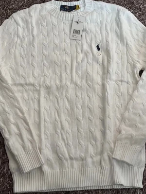 Vit kabelstickad tröja Polo Ralph Lauren - Kabelstickad vit tröja från Polo Ralph Lauren med rund halsringning och ribbade muddar. Klassisk marinblå logga broderad på bröstet. Perfekt för dig som gillar stilrena och tidlösa plagg i garderoben.