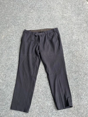 Svarta raka kostymbyxor - Snygga svarta kostymbyxor med rak passform och bälteshällor. Byxorna har två bakfickor med knapp och klassisk dragkedja framtill. Perfekta för en clean och stilren look. Materialet ser ut att vara en blandning av polyester och bomull.