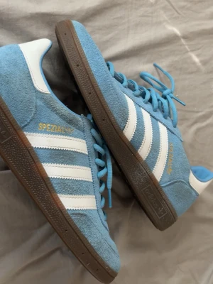 Adidas Spezial blå sneakers - Snygga Adidas Spezial sneakers i ljusblå mocka med klassiska vita ränder och detaljer. Skorna har bruna gummisulor och blå skosnören. Perfekt för dig som gillar att sticka ut lite. Guldfärgad Spezial-logga på sidan ger extra touch. Skorna är nästintill nya, har använt dem 2 gånger men senaste gången råka jag smutsa ner mockan lite o har inte vertygen till att göra rent mockan utan att slita dem. Skrona säljs pågrund av att jag inte använder dem. Hör gärna av dig vid frågor eller funderingar!🤗