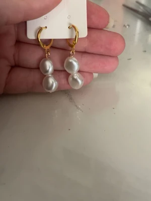 Guldiga örhängen med vita pärlor - Säljer ett par trendiga örhängen med guldfärgade ringar och två vita oregelbundna pärlor på varje örhänge. Perfekt accessoar för dig som gillar en stilren men ändå unik look. Materialet ser ut att vara metall och pärlor.