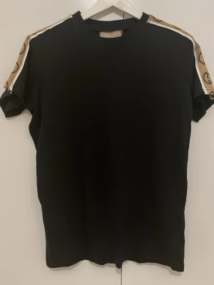 Svart t-shirt med mönstrade axelband - Svart t-shirt med korta ärmar och coola mönstrade band i beige och vitt längs axlarna. Perfekt för en stilren och trendig look. Rund halsringning och rak passform.