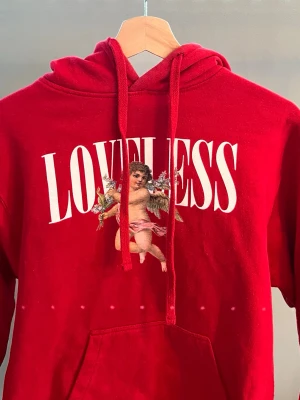 Röd hoodie med LOVLESS tryck - Snygg röd hoodie med stor vit text 'LOVELESS' och en ängel på bröstet. Klassisk känguruficka framtill och justerbar huva med dragsko. Perfekt för dig som gillar streetwear och vill sticka ut lite extra. Mjuk och skön bomullsblandning.