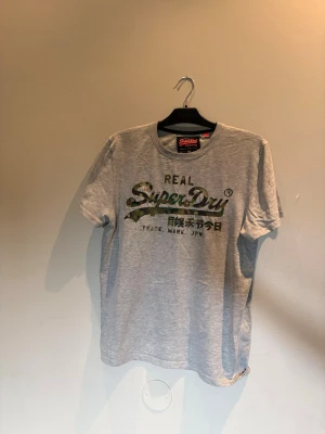 Grå Superdry t-shirt med camologga - Säljer en grå t-shirt från Superdry med stor logga i camomönster och japansk text på bröstet. Klassisk rund hals och korta ärmar. Perfekt för dig som gillar streetwear och vill ha en snygg och bekväm t-shirt.