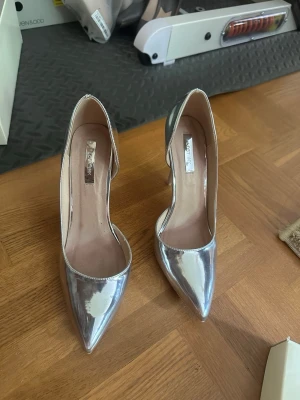 Silvriga spetsiga pumps med hög klack - Säljer ett par glansiga silvriga pumps med spetsig tå och hög smal klack. Skorna har en stilren och modern look med öppen insida och slät finish. Köpte i London använt 3 gånger 