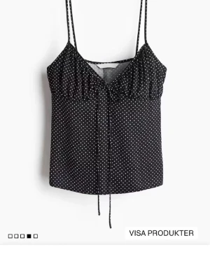 Svart prickig linnetopp med knyt - Snygg svart linnetopp med vita prickar och tunna axelband. Toppen har en söt knyt-detalj framtill och lätt v-ringad hals. Perfekt för varma dagar och enkel att styla med jeans eller kjol.
