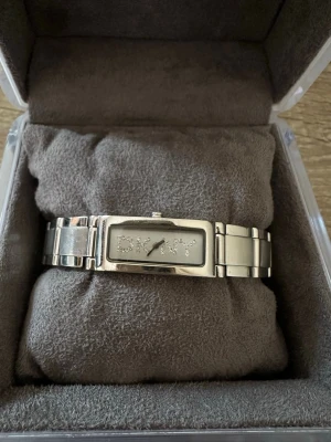 Rektangulär silverfärgad DKNY klocka - Stilren klocka från DKNY i silverfärg med rektangulärt armband och boett. Urtavlan har DKNY-loggan i glittriga stenar och minimalistiska visare. Klockan har en metallänk som ger en modern och trendig look. Inte använd på ett bra tag samt att batteriet behöver bytas ut! OBS: FRAKTAR INOM 24H!🩷