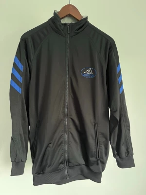 Adidas Track Jacket - Adidas Track Jacket Storlek: XL Passform: Regular Fit Krage: Hög ståkrage Material: Polyester Mått: Bröstvidd 100 cm, längd 74 cm, axel 44 cm Skick: Träningsjackan är i ett väldigt fint skick utan defekter  Nypris cirka: 800 kr