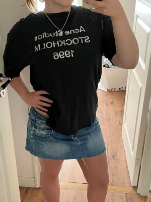Svart Acne Studios t-shirt - Säljer en svart t-shirt med vit text 'Acne Studios Stockholm 1996' på bröstet. T-shirten har normal passform och korta ärmar. Perfekt till jeans eller kjol för en clean och enkel look. Lite smått nopprig