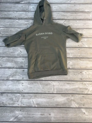 Olivgrön hoodie från Björn Borg - Snygg olivgrön hoodie från Björn Borg med vit text på bröstet där det står 'BJÖRN BORG STOCKHOLM 1956'. Klassisk känguruficka framtill och huva med dragsko. Perfekt för en avslappnad och sportig stil.