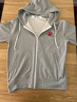 Cdg hoodie - Säljer en grå hoodie med dragkedja och korta ärmar från PLAY. Tröjan har vit dragsko i huvan och ett broderat rött hjärta med ögon på bröstet. Två fickor framtill och mjukt material som känns skönt mot huden. Perfekt för en chill och avslappnad stil.