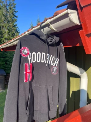 Svart hoodie från Hoodrich med patchar - Svart hoodie från Hoodrich med vita och röda detaljer, stor patchad H på bröstet och tryckta loggor. Klassisk känguruficka framtill och justerbar huva med snörning. Perfekt för dig som gillar streetwear och vill sticka ut.