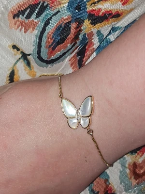 Guldigt armband med pärlemorfjäril - Snyggt armband med tunn guldfärgad kedja och en fjäril i pärlemor-look. Fjärilen har vita och skimrande vingar med guldfärgade kanter och små glittriga stenar i mitten. Perfekt accessoar för dig som gillar unika detaljer och elegant stil.