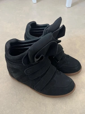 Svarta höga sneakers med kardborre - Säljer ett par svarta sneakers med högt skaft och breda kardborreband. Skorna har en chunky design med kraftig plös och sula i gummi. Materialet är mockaliknande och ger en cool streetwear-look. Perfekta för dig som gillar statement-sneakers.