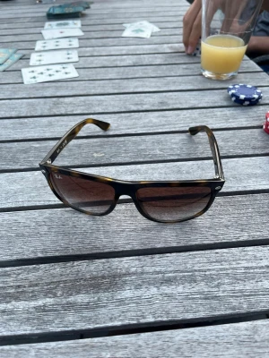 Bruna solglasögon från Ray-Ban - Snygga solglasögon från Ray-Ban med brunt, lätt sköldpaddsmönstrat båge och rektangulär form. Glasen är tonade i brunt och bågarna har diskreta metallnitar vid fronten. Perfekta för soliga dagar.