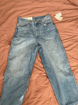 Ljusblå raka jeans med hög midja - Säljer ett par ljusblå jeans med rak passform och hög midja. Klassisk femficksmodell med knappgylf och bälteshällor. Jeansen har en avslappnad look och är tillverkade i denim. Perfekta för en trendig och enkel stil.