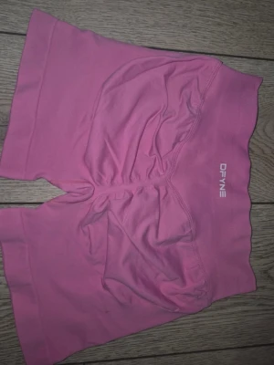Rosa träningstights shorts från DFYNE - Säljer ett par rosa träningstights shorts från DFYNE. De har hög midja, tight passform och är tillverkade i ett stretchigt syntetmaterial. Perfekta för gymmet eller annan träning. Logga på linningen framtill.