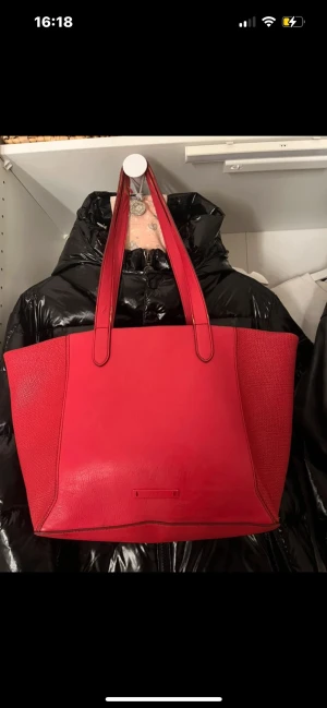 Röd shoppingväska i skinnimitation - Snygg röd shoppingväska med långa axelband och rymlig insida. Väskan har slät framsida och baksida medan sidorna har en strukturerad, vävd look. Tillverkad i skinnimitation och perfekt för dig som vill ha plats för allt du behöver under dagen.