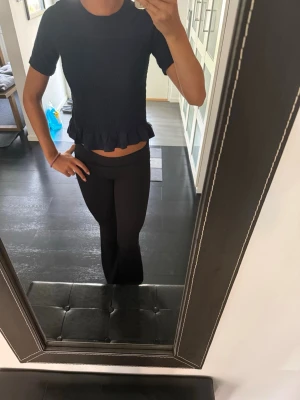 Svart topp med volangkant - Säljer en mörk blå topp med korta ärmar och rund hals. Toppen har en ribbad struktur över magen och avslutas med en söt volangkant nedtill. Perfekt för dig som gillar en enkel men ändå detaljrik stil.