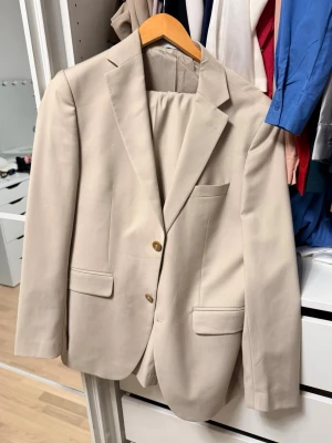 Beige kostym med kavaj och byxor - Stilren beige kostym med klassisk kavaj och matchande byxor. Kavajen har två knappar, två fickor med lock och en bröstficka. Byxorna har raka ben och bälteshällor. Perfekt för dig som vill ha en clean och modern look. Stl: 48 typ en medium