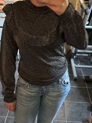 Glittrig grå tröja med volang Isolde - Mysig gråmelerad tröja från Isolde med glittrigt material och volangdetalj över axlar och rygg. Långärmad modell med rund halsringning och stretchig passform. Perfekt för dig som gillar en trendig och bekväm look. I storlek L.