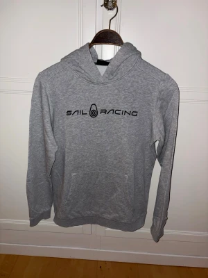 Grå hoodie Sail Racing - Snygg grå hoodie från Sail Racing med svart logga på bröstet och en mindre logga på huvan. Klassisk känguruficka framtill och mjukt material, perfekt för en avslappnad och sportig stil. Storlek 164-176