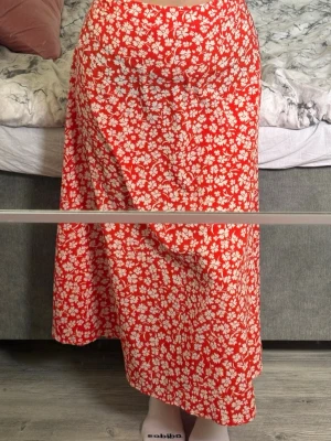 Röd blommig långkjol - Säljer en röd/orange långkjol med vitt blommönster. Kjolen har en flowy passform och är perfekt för varmare dagar. Materialet känns lätt och bekvämt mot huden. Supersnygg att styla med en enkel topp eller stickad tröja. Har en dragkejda på baksidan. 
