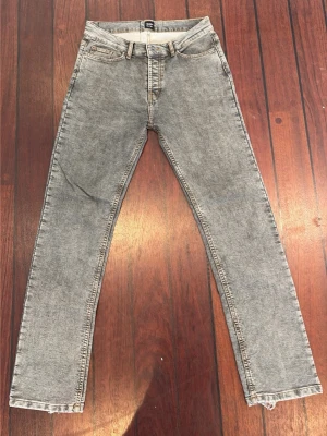 Grå tvättade jeans från Montén - Säljer ett par grå tvättade jeans från Montén med klassisk femficksmodell och raka ben. Jeansen har kontrastsömmar och en stor grön patch bak med texten 'Classic Way of Life'. Perfekta för dig som gillar en avslappnad och trendig stil.